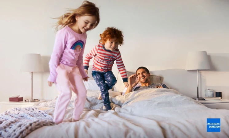 Two children jumping on a bed with their dad smiling in the background, illuminated by lamps on the nightstands in a home setting.