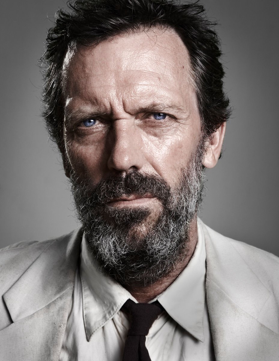 Close-up portrait of an adult male with graying hair and beard, wearing a light-colored suit and dark tie against a gray background.