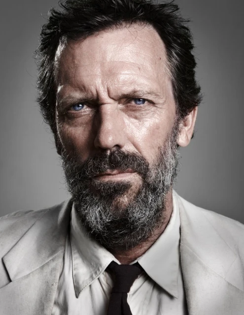 Close-up portrait of an adult male with graying hair and beard, wearing a light-colored suit and dark tie against a gray background.