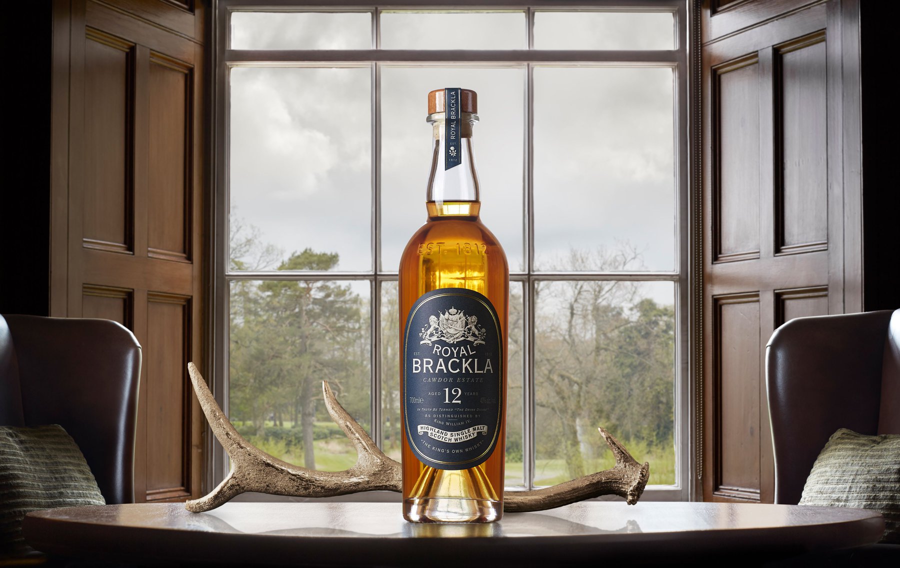 A bottle of Royal Brackla whisky sits on a wooden table in front of an open window, flanked by chairs and a set of antlers.