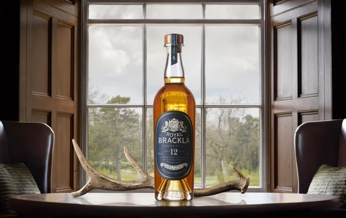 A bottle of Royal Brackla whisky sits on a wooden table in front of an open window, flanked by chairs and a set of antlers.