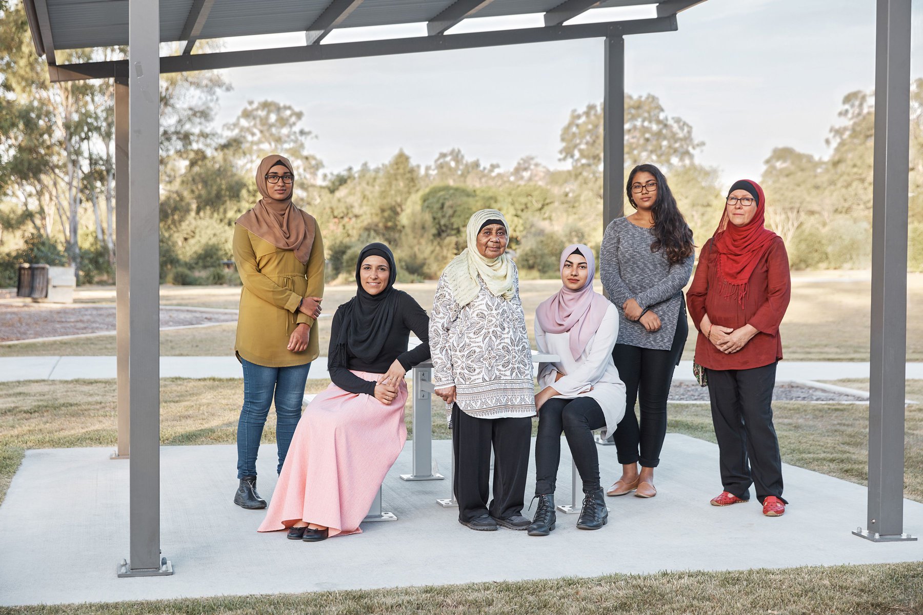 Group of six women in hijabs outdoors. Various colors and styles, seated and standing in a park-like setting. Neutral daytime lighting.