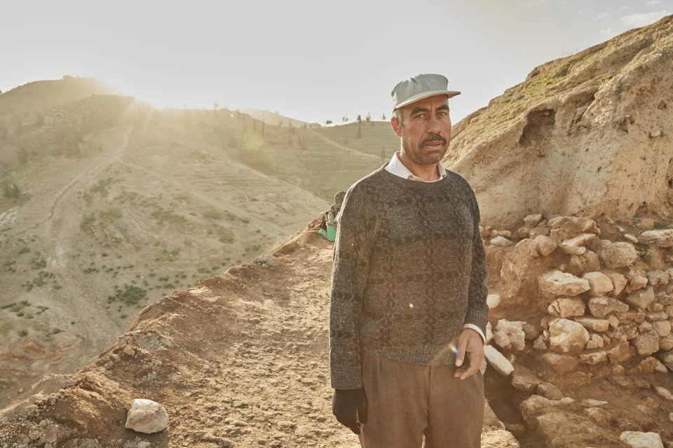 Portrait of a middle-aged man in a cap standing on a dirt path with terraced hills and bright sunlight in the background.  