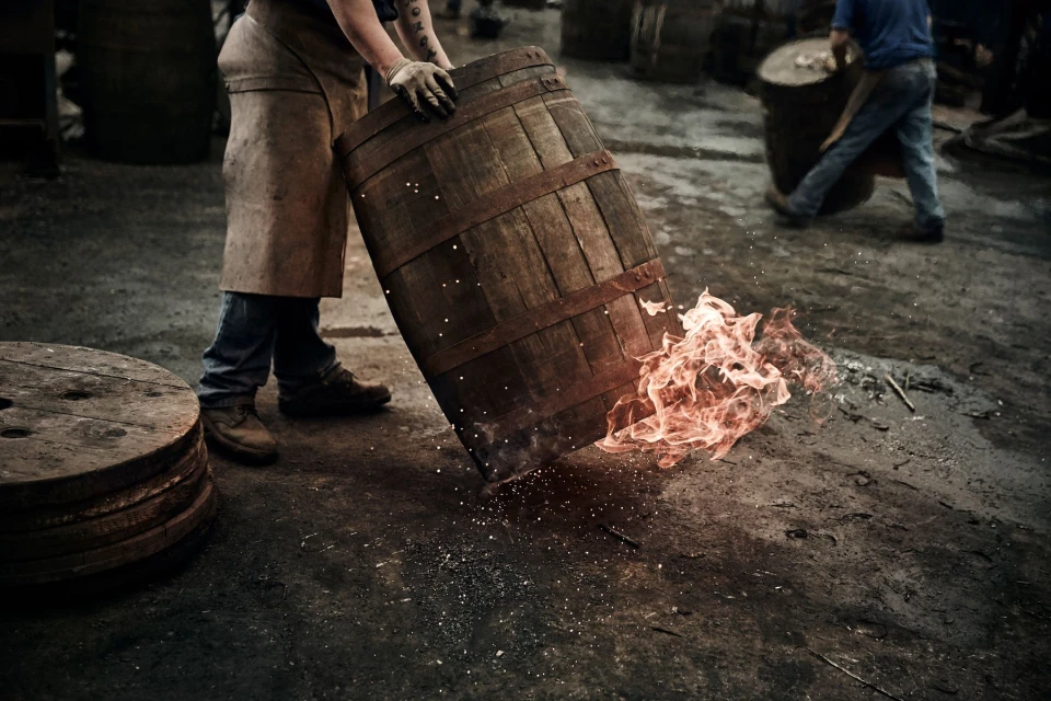 Craftsman in a brown apron and gloves holds a wooden barrel with flames erupting in a dimly lit workshop, barrel making industry.