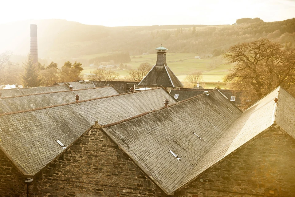 Exterior view of a stone distillery with multiple gabled roofs and a chimney against a hillside in golden light.