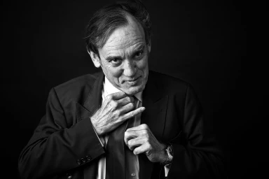 Monochrome portrait of an older man adjusting his tie while wearing a dark suit against a dark background