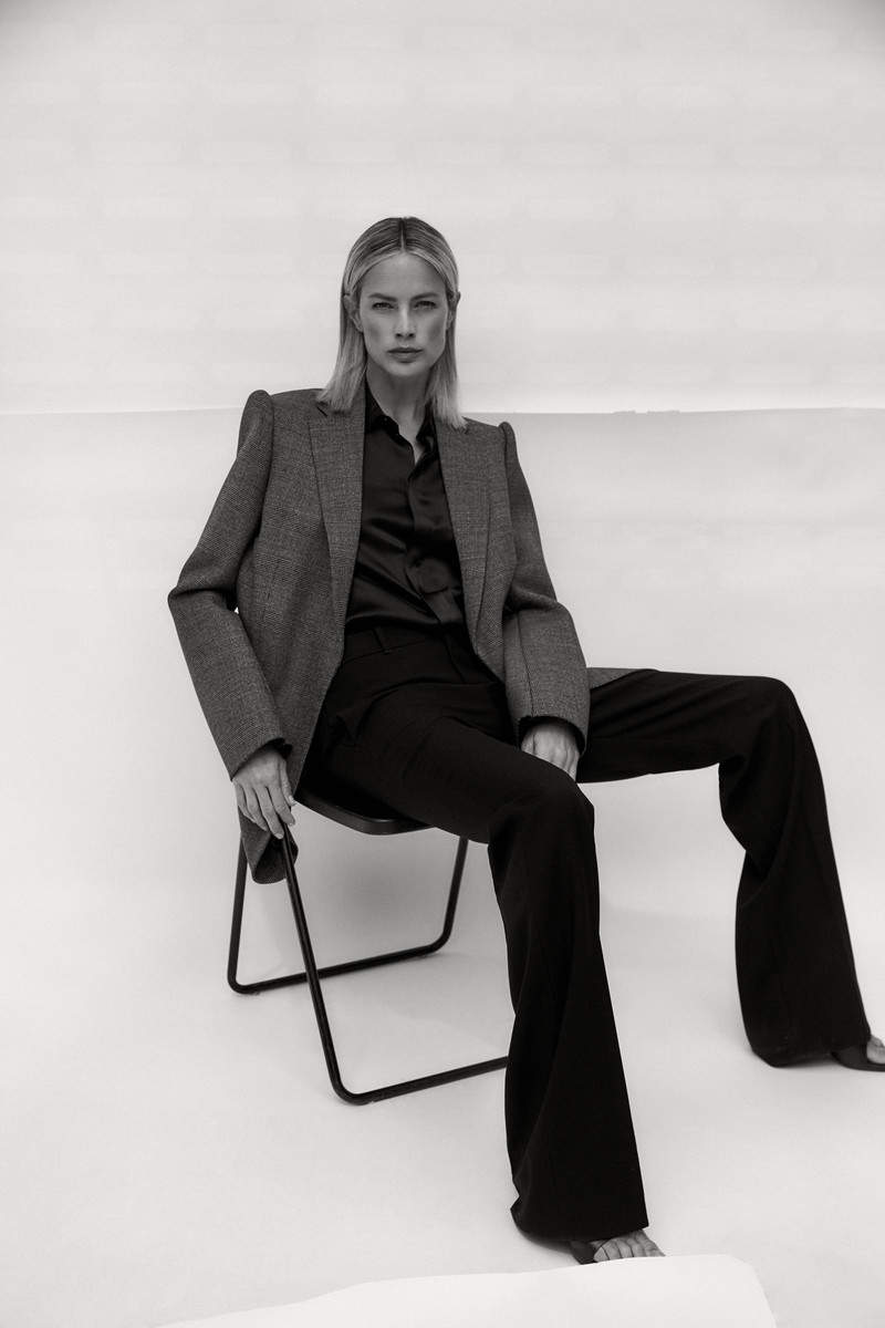 Monochrome fashion portrait of a blonde adult woman in a blazer and bell-bottom pants, seated on a chair against a white backdrop.