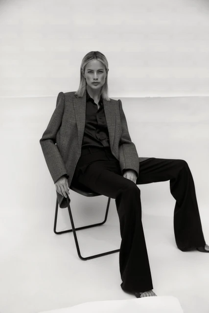 Monochrome fashion portrait of a blonde adult woman in a blazer and bell-bottom pants, seated on a chair against a white backdrop.