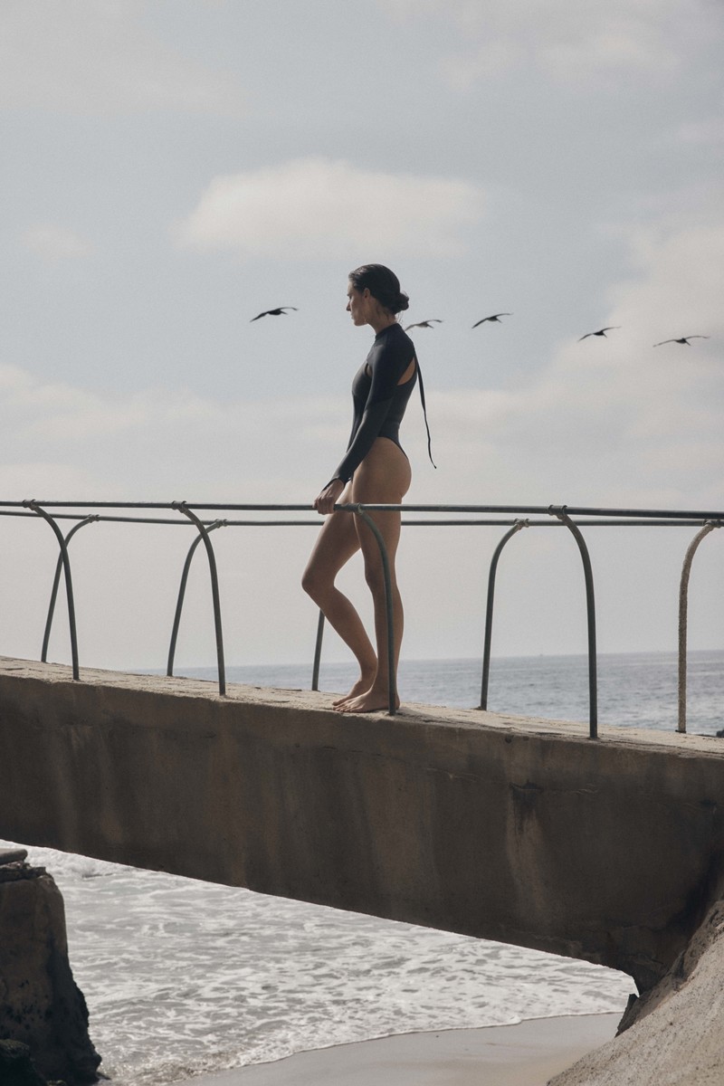 Woman in a black bodysuit walks along a concrete bridge near a sandy beach with waves and birds flying overhead on a cloudy day.