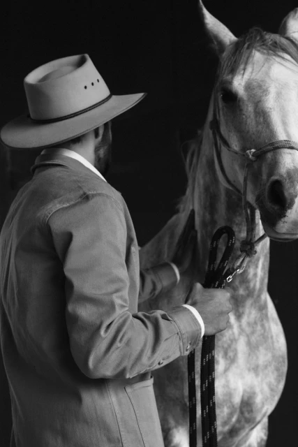 Monochrome portrait of a horse and a man in a cowboy hat and jacket holding the horse's lead against a dark background.