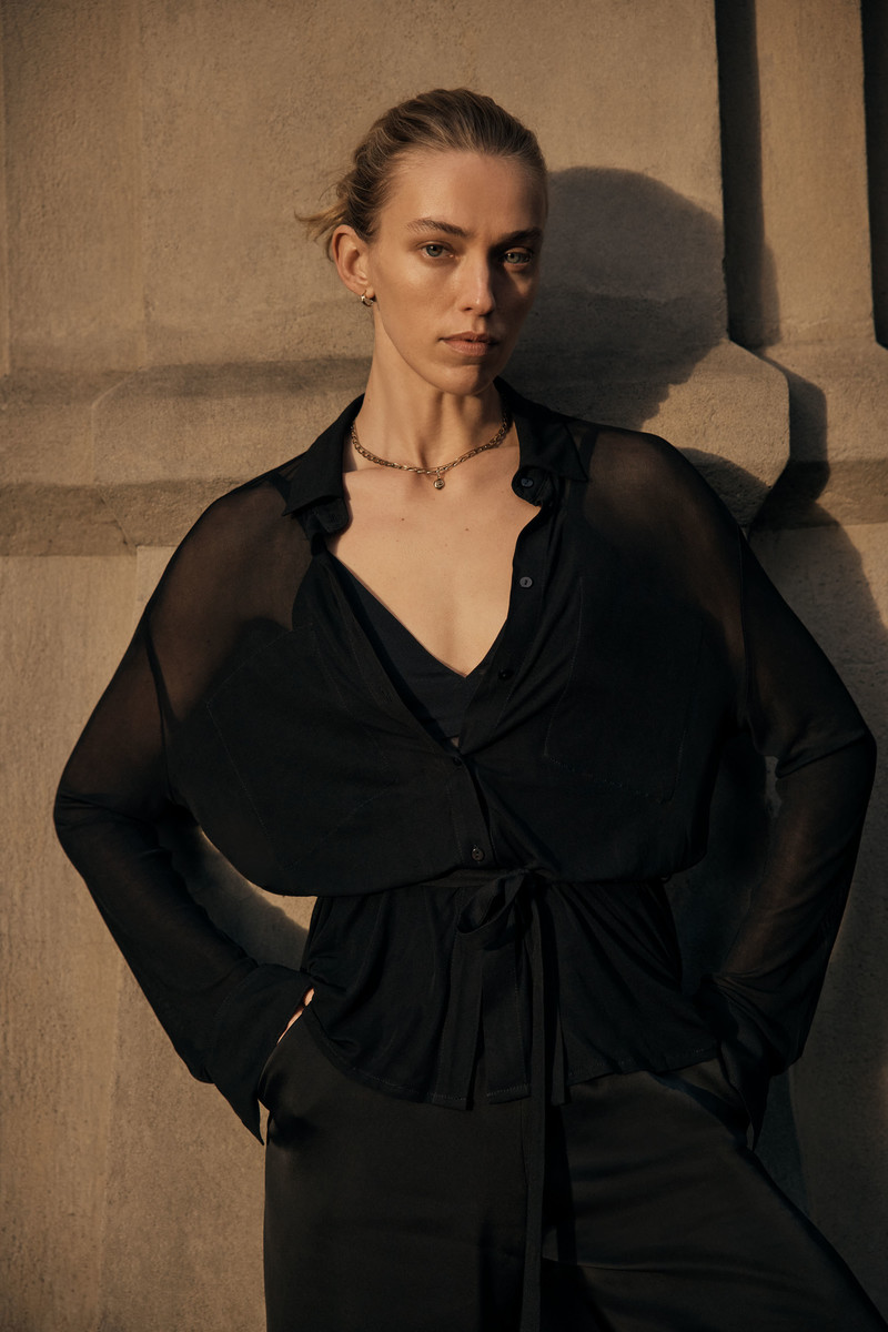 An adult woman stands indoors wearing a black blouse and pants with concrete columns in the background, bathed in soft, warm light.