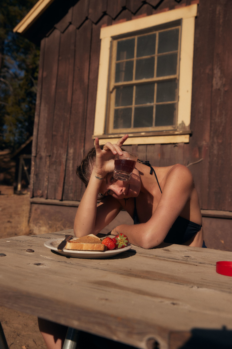 A woman in a black bikini top sitting at a picnic table with toast, strawberries, and a drink next to a rustic cabin.