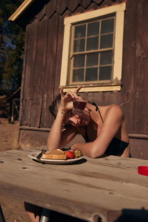 A woman in a black bikini top sitting at a picnic table with toast, strawberries, and a drink next to a rustic cabin.
