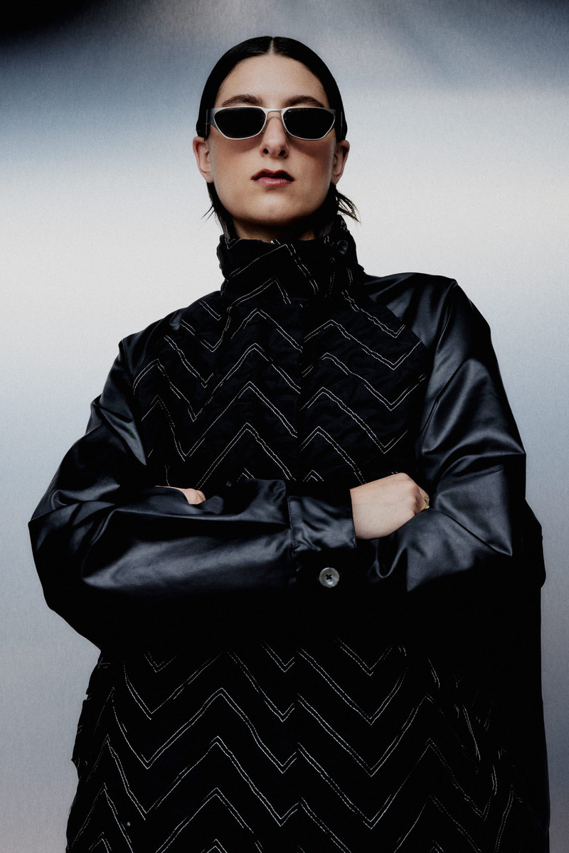 Fashion portrait of a woman in a black jacket with white chevron patterns, wearing sunglasses, set against a blurred grey background.