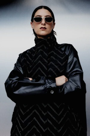 Fashion portrait of a woman in a black jacket with white chevron patterns, wearing sunglasses, set against a blurred grey background.