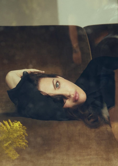 A woman with long dark hair reclines on a brown couch, eyes gazing directly at the viewer. The shot is a medium close-up, with indoor lighting.