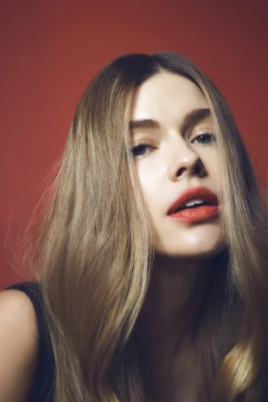 Close-up portrait of a woman with long blonde hair and red lipstick against a reddish-brown background.