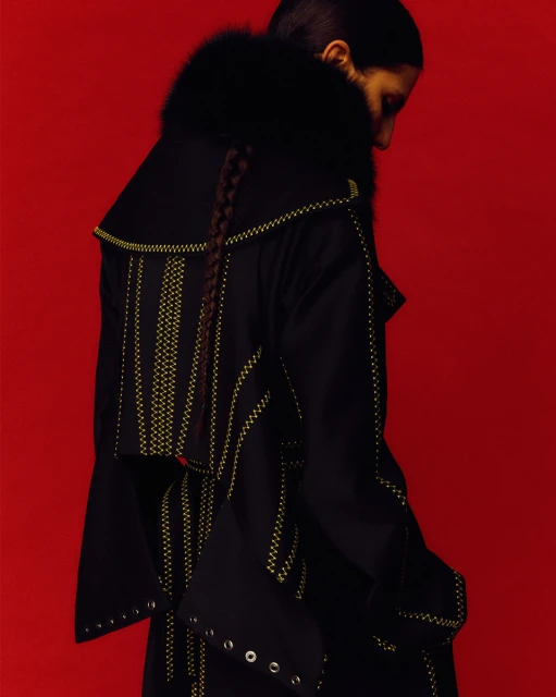 Fashion shot of a woman in a black coat with fur collar and decorative stitching, set against a vivid red background in studio lighting.