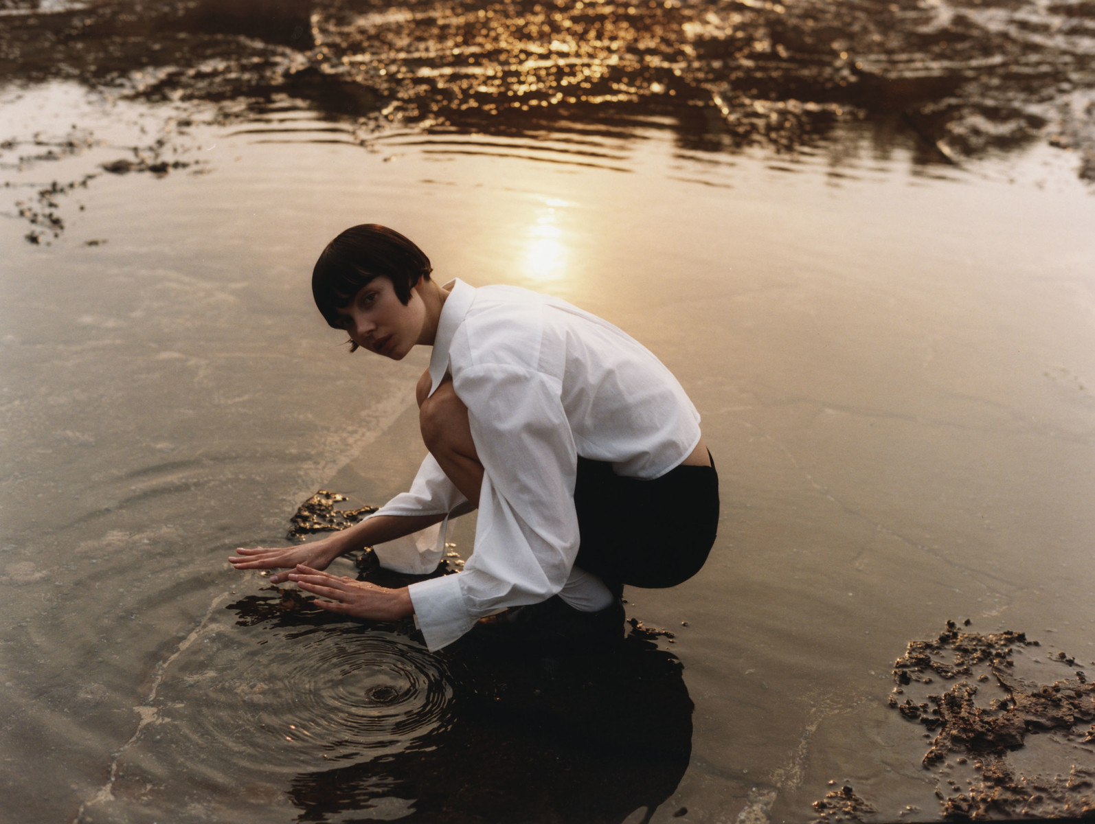 Woman in white shirt crouching and touching rippling water at sunset by photographer Darren Mcdonald