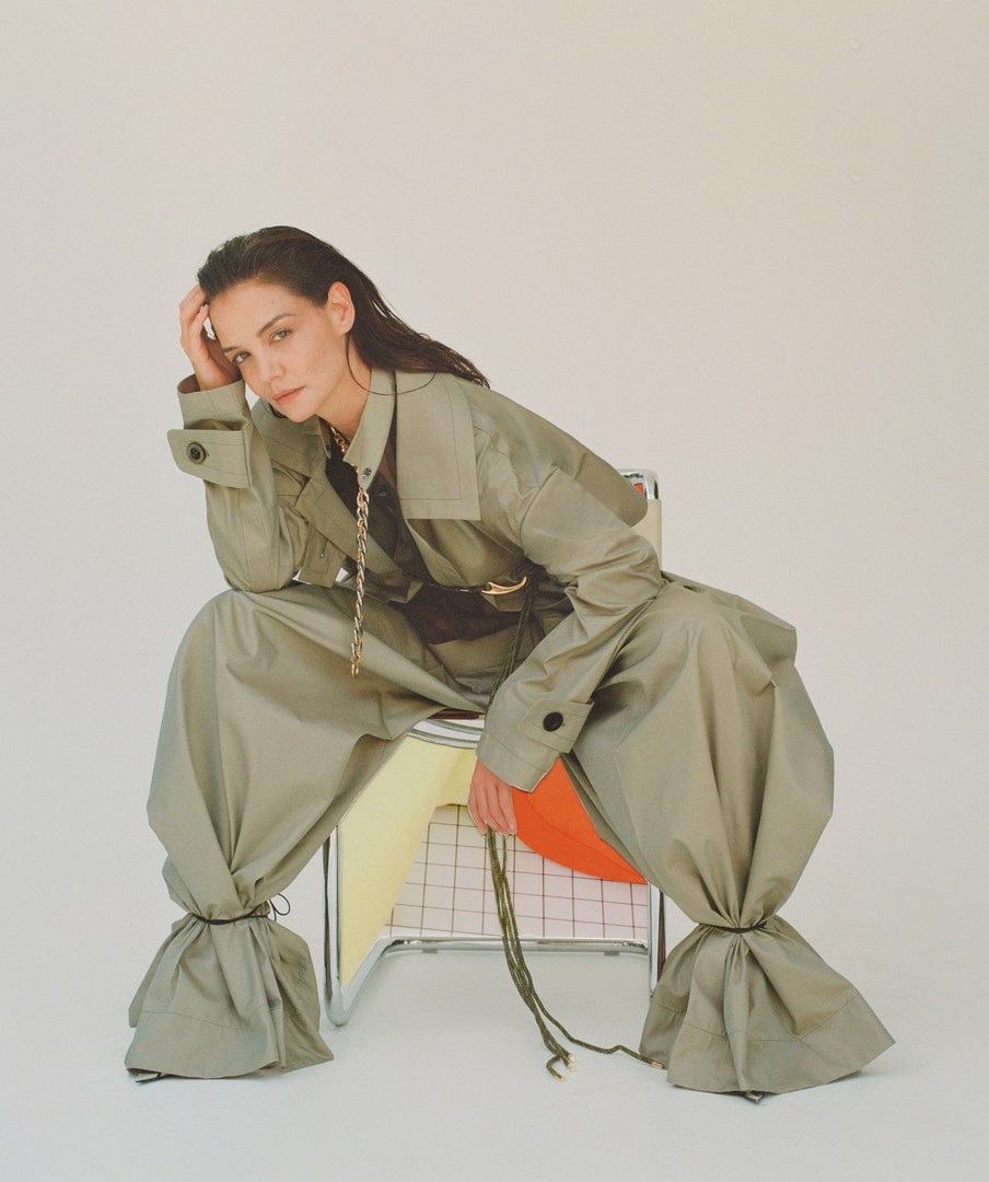 Woman in an olive-green coat and puffy pants sitting on a chair against a white background. The image is a fashion editorial.