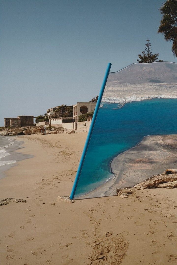 Beach scene with blue pipe sticking out of sand, mirroring a print of ocean view, houses on the shore, clear blue sky, sunny day.
