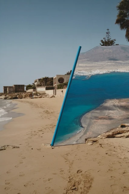 Beach scene with blue pipe sticking out of sand, mirroring a print of ocean view, houses on the shore, clear blue sky, sunny day.