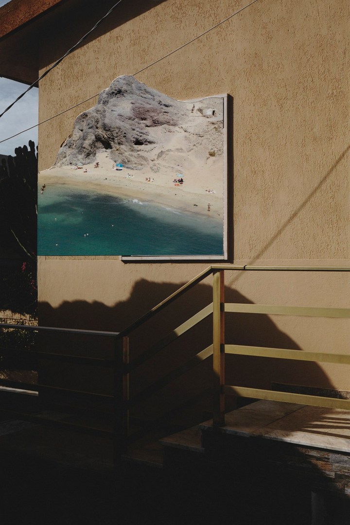 Framed photograph of a beach scene hanging on the exterior of a tan building with railing shadows, creating an urban juxtaposition.