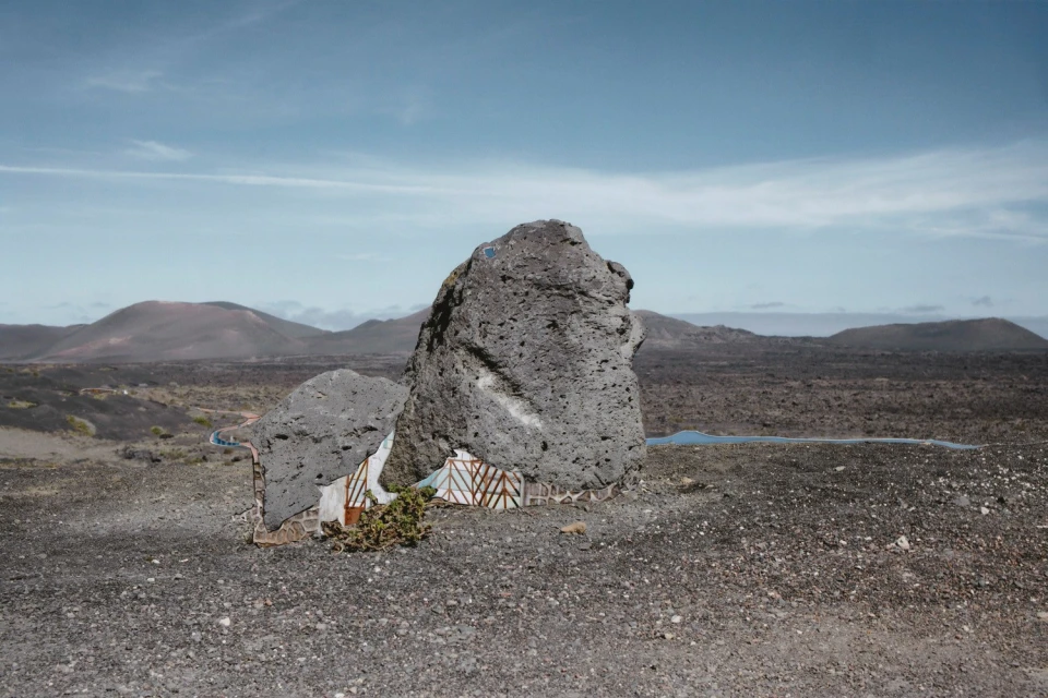 Volcanic landscape with rock formation, shrubs, and distant mountains under a blue sky with some clouds, creating a scenic vista.