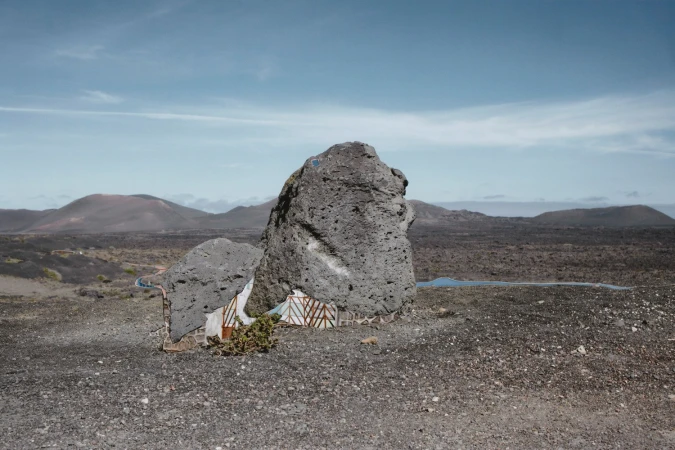 Volcanic landscape with rock formation, shrubs, and distant mountains under a blue sky with some clouds, creating a scenic vista.