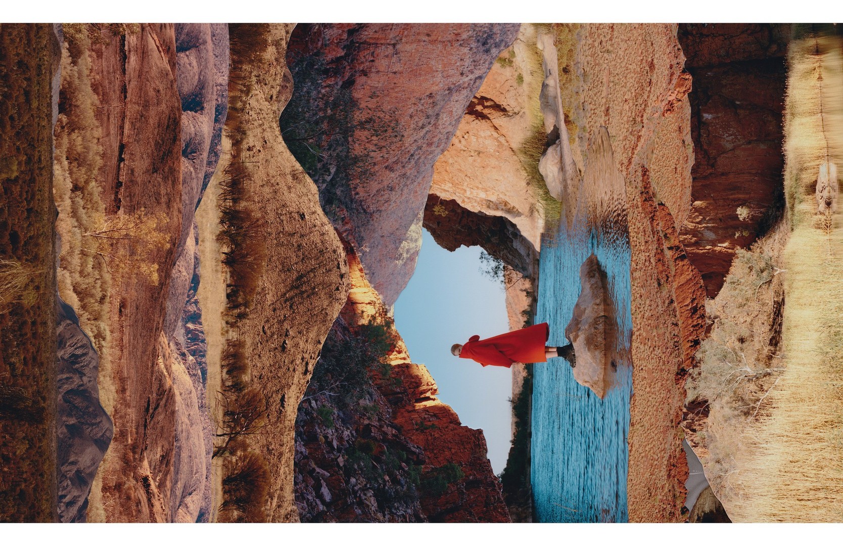 A person in a red robe stands on a rock in a river surrounded by canyon walls and blue sky, shot with a wide-angle lens in an outdoor setting.