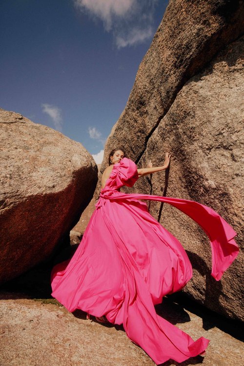 A woman in a pink gown stands next to a large rock formation under a blue sky, creating a striking contrast of color and form.