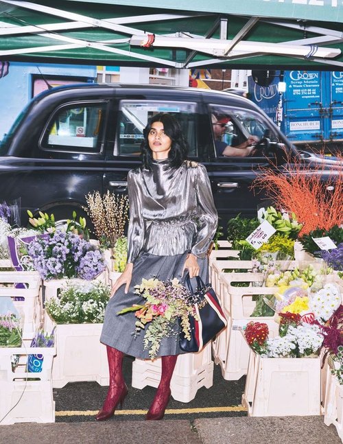Woman poses at flower market in metallic top, gray skirt, and red boots, holding floral bouquet and handbag in outdoor setting.