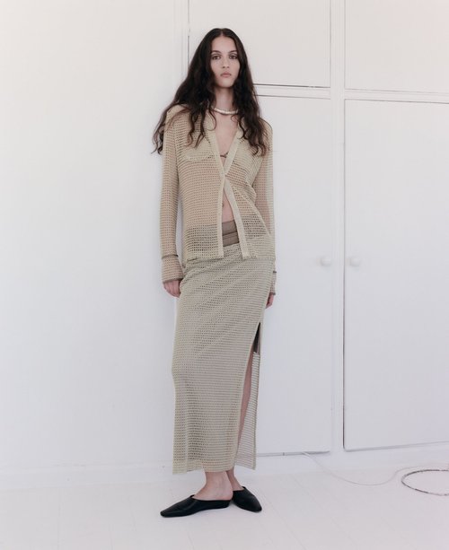 Indoor shot of an adult female model wearing a sheer knit skirt and blouse with black flats against a white wall, full-length portrait.
