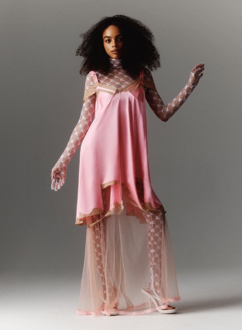 A fashion shot of a slender adult female model with dark curly hair wearing a pink lace-trimmed slip dress and patterned top and tights.