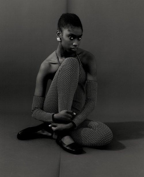 Black and white studio portrait of a woman with short hair, wearing a shimmering jumpsuit and flats, sitting cross-legged on a gray background.