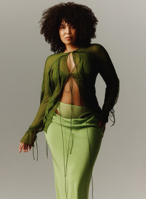 Portrait of an adult woman with an afro, wearing a green sheer blouse and skirt, posing in a photo studio with a grey backdrop.