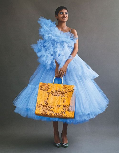 A stylish adult woman in a light blue tulle dress holding a tan patterned handbag, posing in a studio against a grey backdrop.