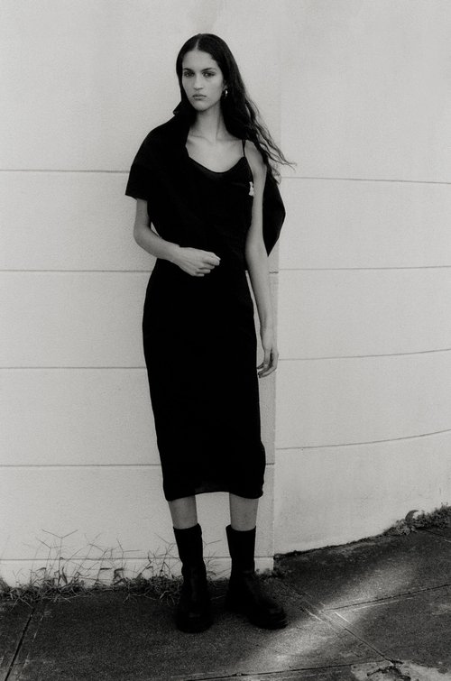 A woman in a black dress and boots stands against a textured wall in a full-length shot, showcasing fashion and style in monochrome.