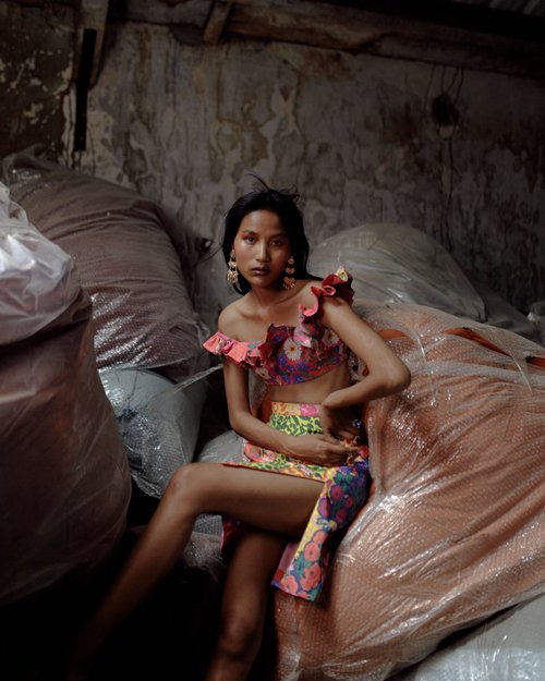 Adult female model with dark hair sits among plastic-wrapped packages, wearing a floral two-piece outfit and gold earrings in an indoor setting.