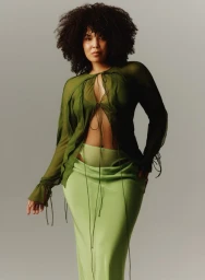 Woman with dark curly hair wearing a sheer olive green tie-front top and a light green maxi skirt against a grey studio backdrop.