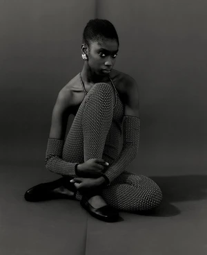 Black woman seated on a studio floor wearing a patterned bodysuit with separate arm sleeves and dark flats, styled by Ria Kamat Shetty.