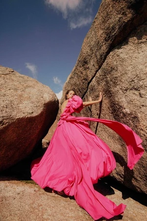 Model in voluminous bright pink gown posing between massive weathered granite boulders under clear sky