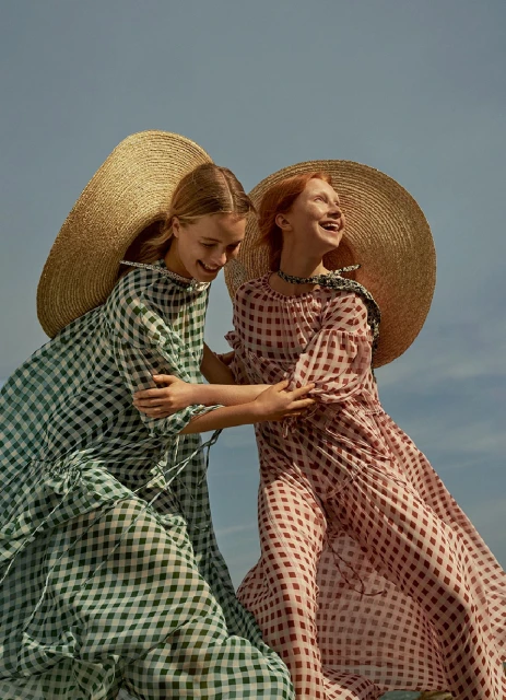 Two laughing girls wearing oversized straw hats and checkered dresses embrace against a light blue sky in a fashion outdoor shot.