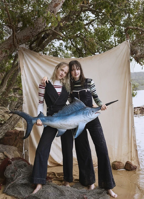 Two women pose with a large, blue marlin on a beach with a backdrop of trees, sand, and water; wearing striped tops and dark pants.