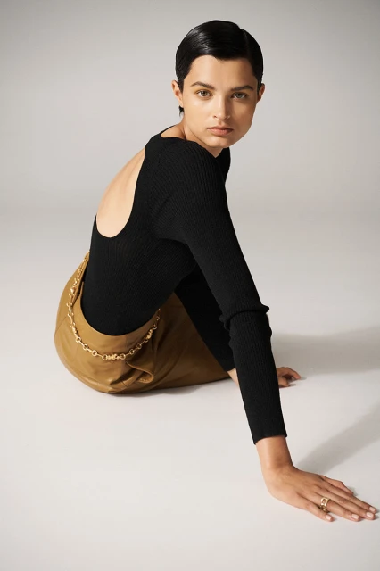 Fashion shot of a woman with short dark hair wearing a black long-sleeved shirt and a tan skirt posing in a photo studio with a light background.