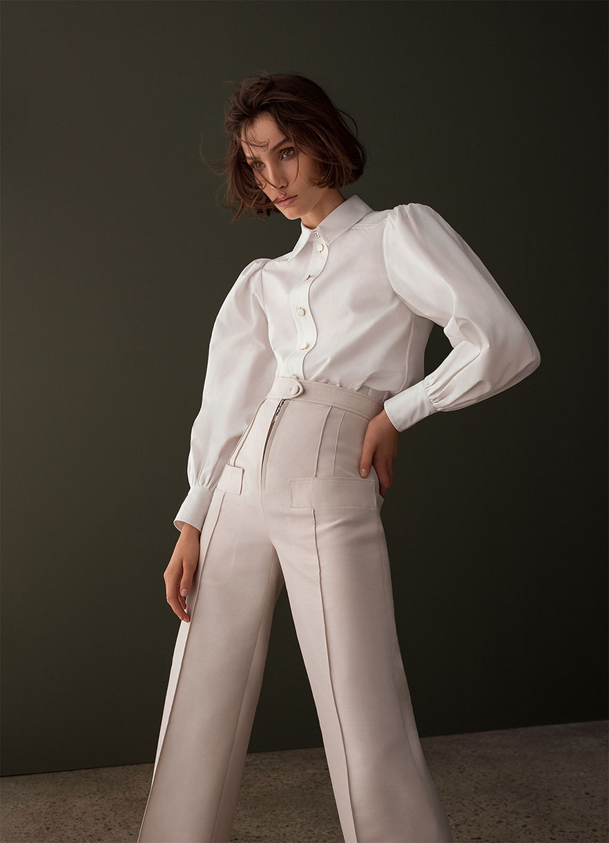 Woman in white blouse and cream-colored trousers poses against a dark green background with a semi-glossy texture, casting soft studio lighting.