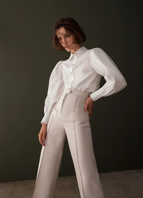 Woman in white blouse and cream-colored trousers poses against a dark green background with a semi-glossy texture, casting soft studio lighting.