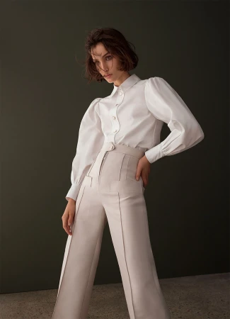 Woman in white blouse and cream-colored trousers poses against a dark green background with a semi-glossy texture, casting soft studio lighting.