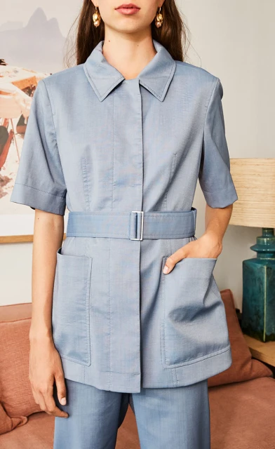 A model wears a light blue collared short-sleeve belted jacket with pockets and matching pants in an indoor setting with a neutral color palette.