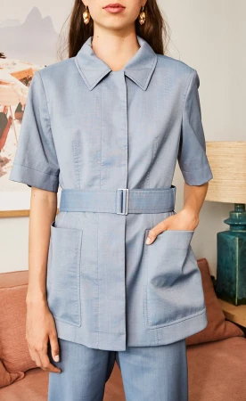 A model wears a light blue collared short-sleeve belted jacket with pockets and matching pants in an indoor setting with a neutral color palette.